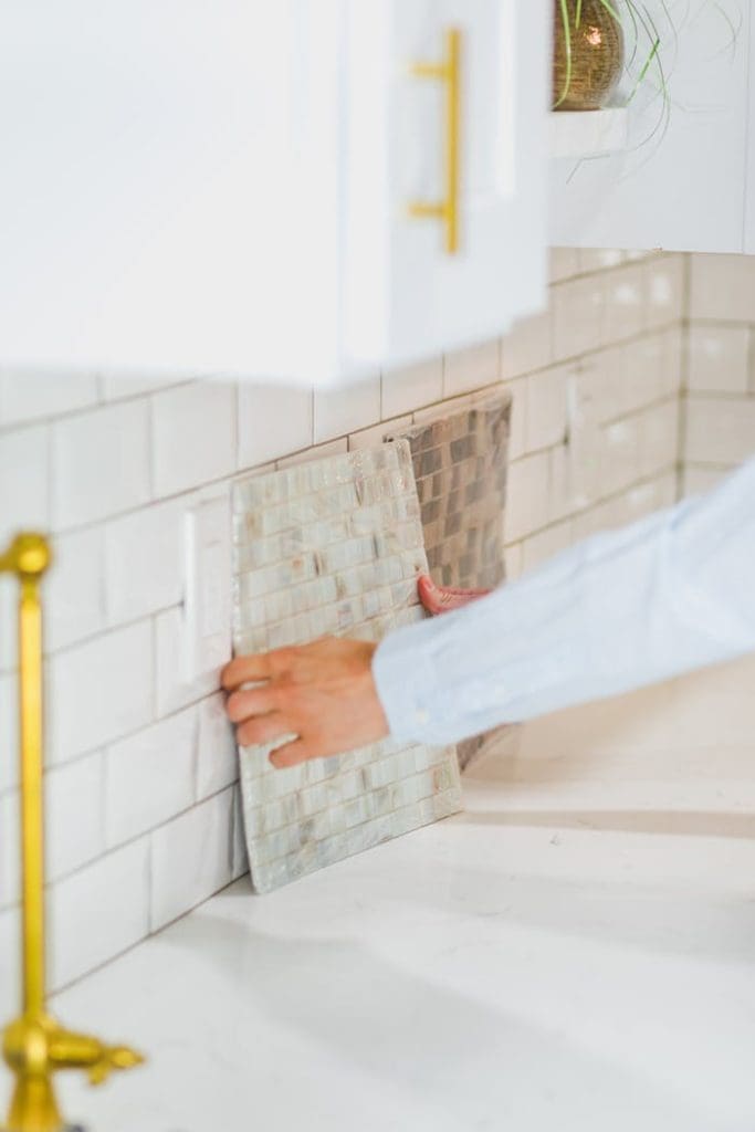 person-choosing-a-tile-design-on-wall-8293686 Person installing tile backsplash in a bright, modern kitchen setting.